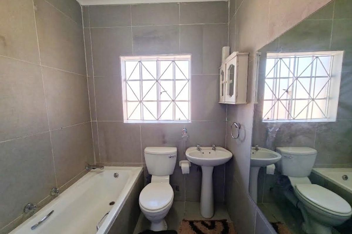 A functional bathroom featuring a bathtub, pedestal sink, toilet, and large wall mirror, finished with neutral wall tiles and security-barred windows.