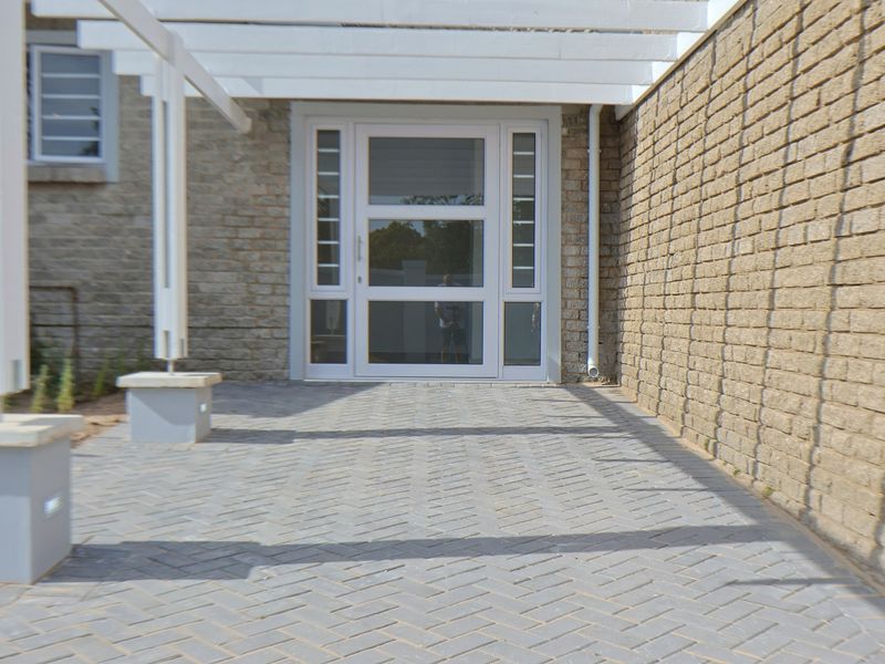 A well-maintained front entrance featuring a modern glass-panelled door, stone-clad walls, and a paved patio area sheltered by a white pergola.