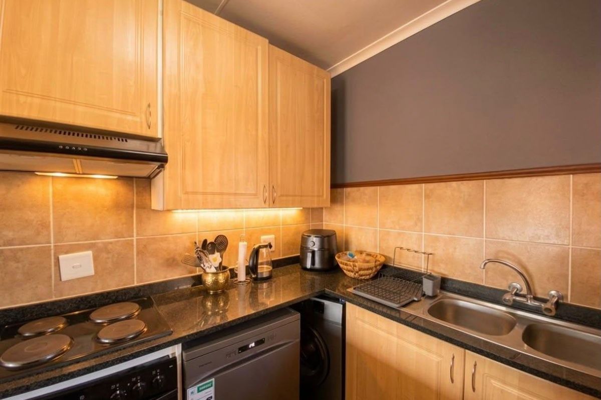 Compact kitchen featuring light wood cabinetry, granite countertops, a double stainless steel sink, and a tiled backsplash with integrated under-cabinet lighting.