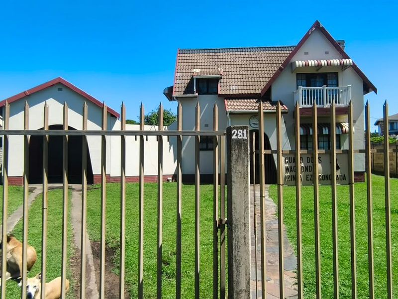 Front view of a two-story house featuring a tiled roof, balcony, and detached garage, enclosed by metal perimeter fencing and a paved walkway.