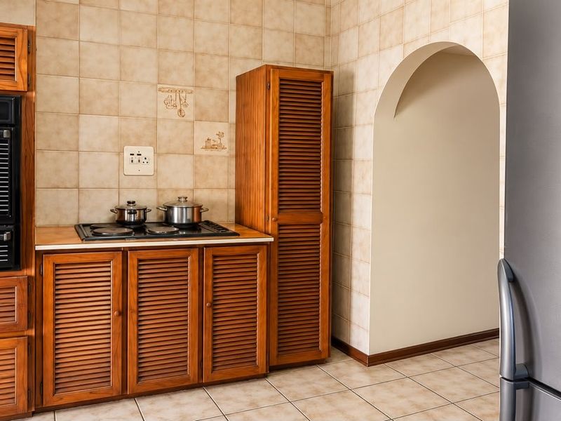 A functional kitchen featuring traditional louvered wooden cabinetry, a built-in oven, hob, and neutral tiled finishes with an arched entryway.