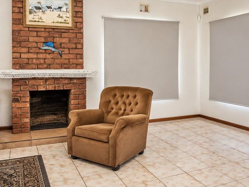 A living room featuring a classic brick fireplace, neutral tiled flooring, and fitted roller blinds, offering a functional and well-lit space.
