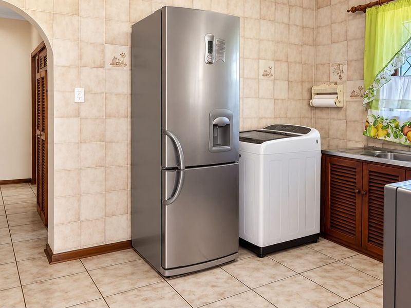A functional laundry area featuring tiled walls and flooring, built-in wooden cabinetry, and a stainless steel sink with a window view.