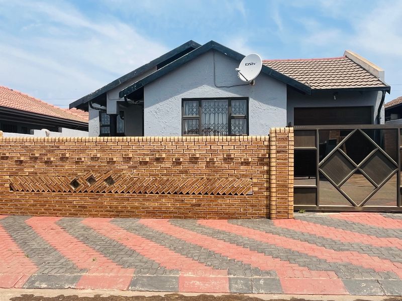 Front view of a residential house featuring a decorative brick boundary wall, metal gate, paved driveway, and tiled roof.