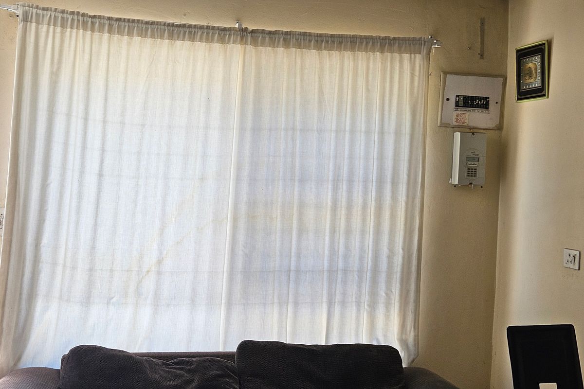 A living area featuring a large window with curtain rail, wall-mounted electrical distribution board, and alarm system keypad.