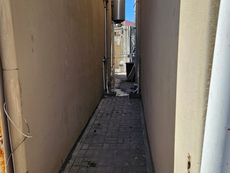 A narrow side passage between townhouse walls featuring paved flooring, external plumbing, and a wall-mounted geyser unit.