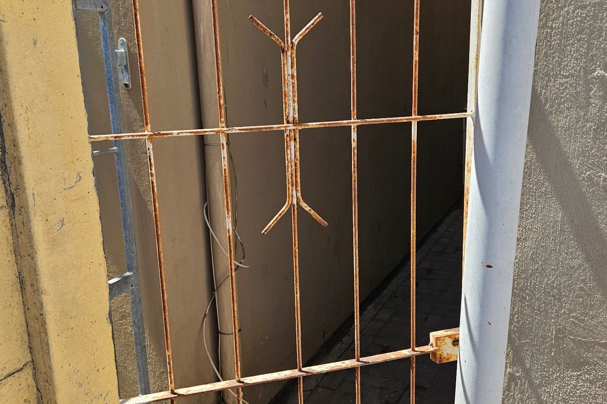 A narrow side passage featuring a rusted metal security gate, paved walkway, and exterior wall with visible paint wear.
