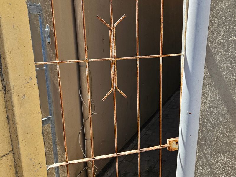 A narrow side passage featuring a rusted metal security gate, paved walkway, and exterior wall with visible paint wear.