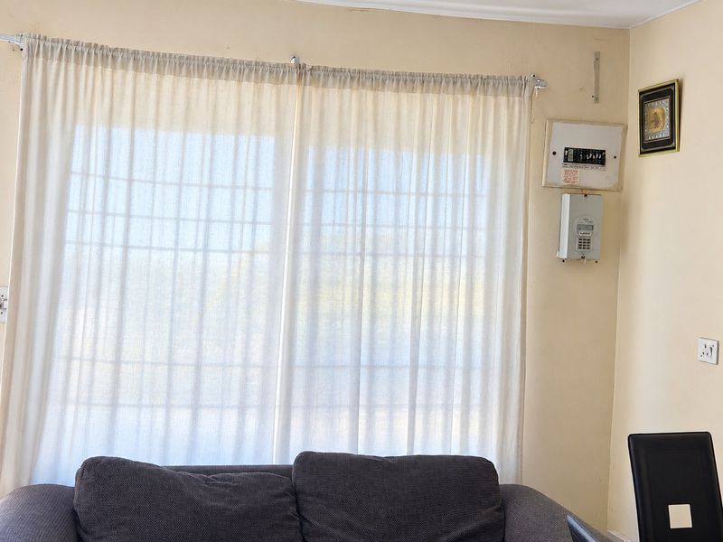 A living room area featuring a large window with curtains, an electrical distribution board, and a prepaid electricity meter mounted on the wall.