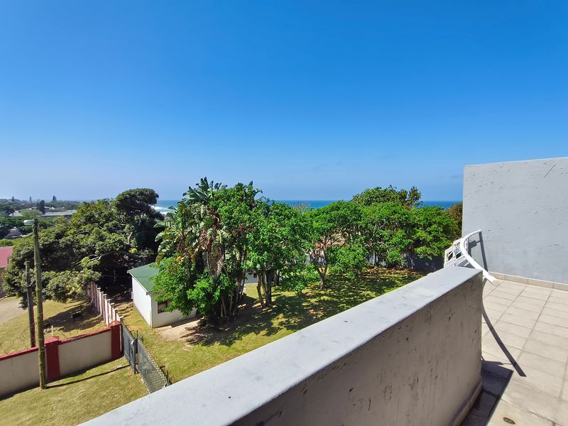A spacious balcony featuring tiled flooring and a concrete balustrade, offering an elevated view of the surrounding garden and distant ocean.