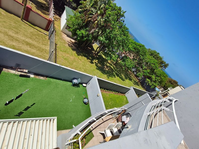 An elevated aerial view showing the apartment balcony, private artificial grass area, and surrounding landscaped grounds with coastal greenery.