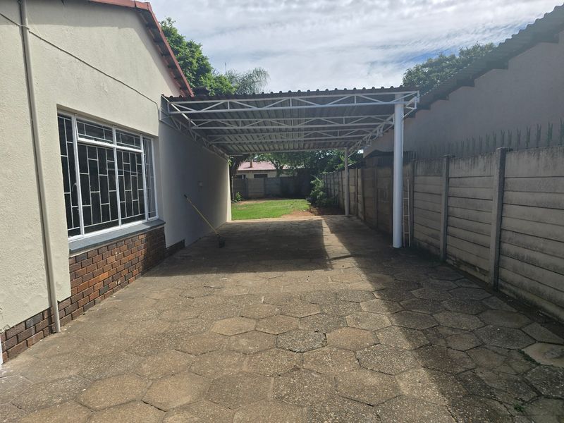 A paved rear driveway featuring a metal carport structure, enclosed by precast concrete boundary walls and a brick-plinth house exterior.