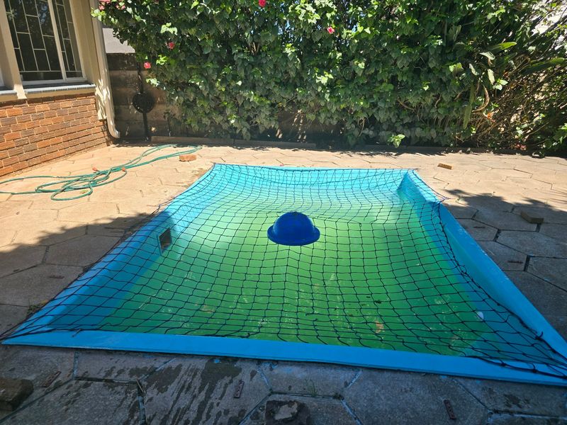 Outdoor swimming pool area featuring a safety net and paved surround, currently requiring maintenance to the pool water and surrounding paving.