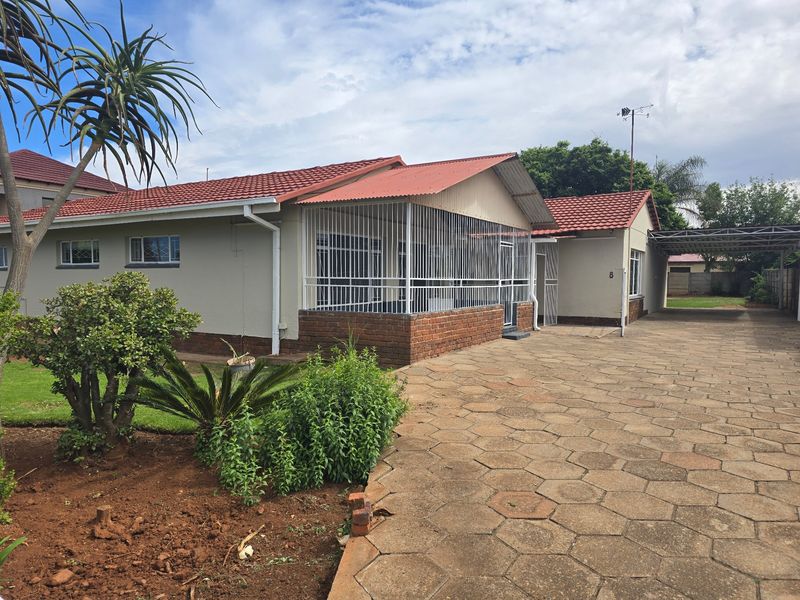 Front view of a single-story house featuring a tiled roof, brick accents, security bars, and a spacious paved driveway with a carport.
