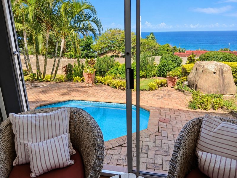 Outdoor pool area featuring a brick-paved patio, swimming pool, and landscaped garden with ocean views, accessed via a sliding glass door.