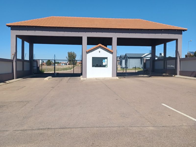 A well-maintained residential estate entrance featuring a covered security gatehouse, asphalt driveway, and perimeter fencing under a clear blue sky.