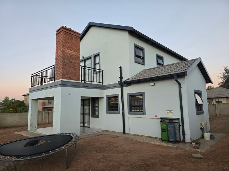 Rear view of a two-story house featuring a brick chimney, upper-level balcony, and paved patio area with security fixtures.