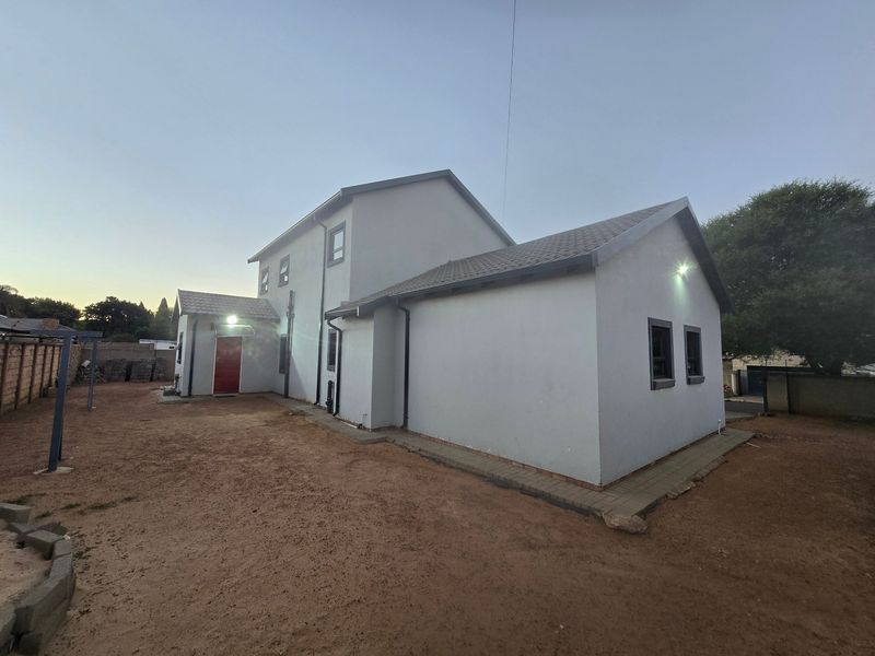Rear view of a two-story house featuring plastered walls, a tiled roof, and security lighting, set within an unlandscaped yard with a perimeter boundary wall.