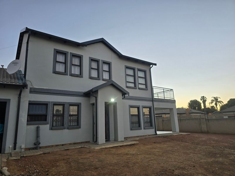 A two-story residential exterior featuring a balcony, covered patio, and security bars on windows, set against a large, unlandscaped rear yard.