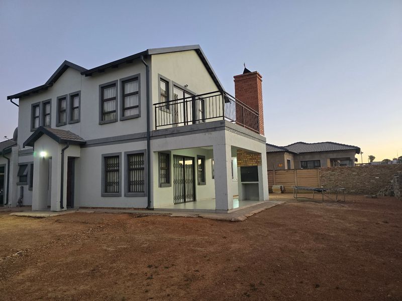 Rear view of a double-story house featuring a balcony, built-in braai area, and a spacious backyard with potential for landscaping.