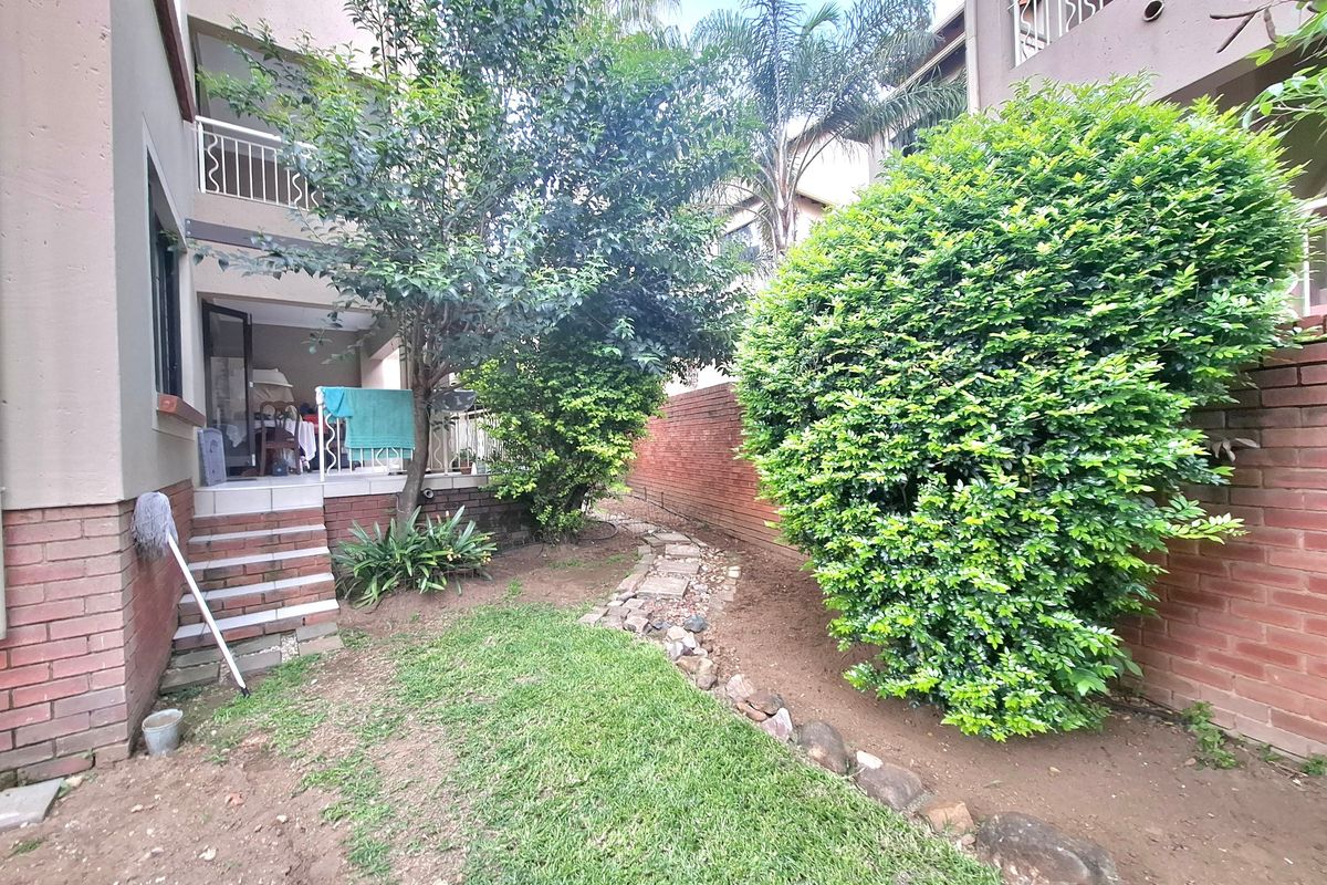 A rear garden area featuring a brick boundary wall, landscaped greenery, and tiled steps leading up to a covered patio balcony.