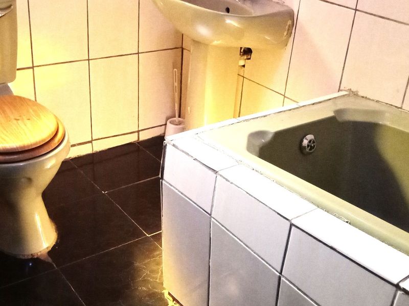 A dated bathroom featuring a pedestal basin, toilet, and tiled bathtub with dark floor tiles and light wall tiles.