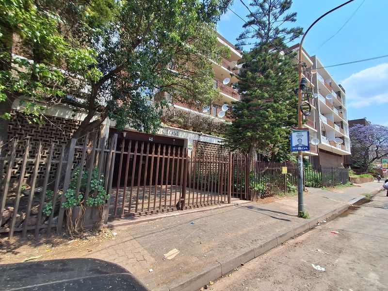 Street view of the apartment building facade featuring a secure gated entrance, multi-level balconies, and established trees along the sidewalk.