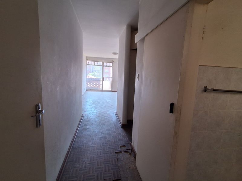 A vacant hallway featuring parquet flooring with visible damage and missing sections, leading towards an open-plan living area with balcony access.
