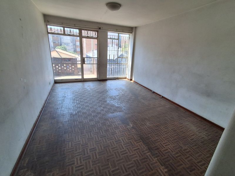 An empty living room featuring original parquet flooring, large windows with security bars, and neutral walls in need of cosmetic refurbishment.