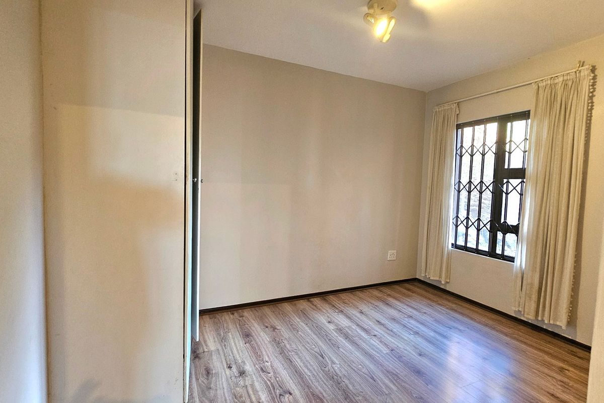 A bright, empty bedroom featuring modern laminate flooring, a built-in cupboard, and a window fitted with a security gate.