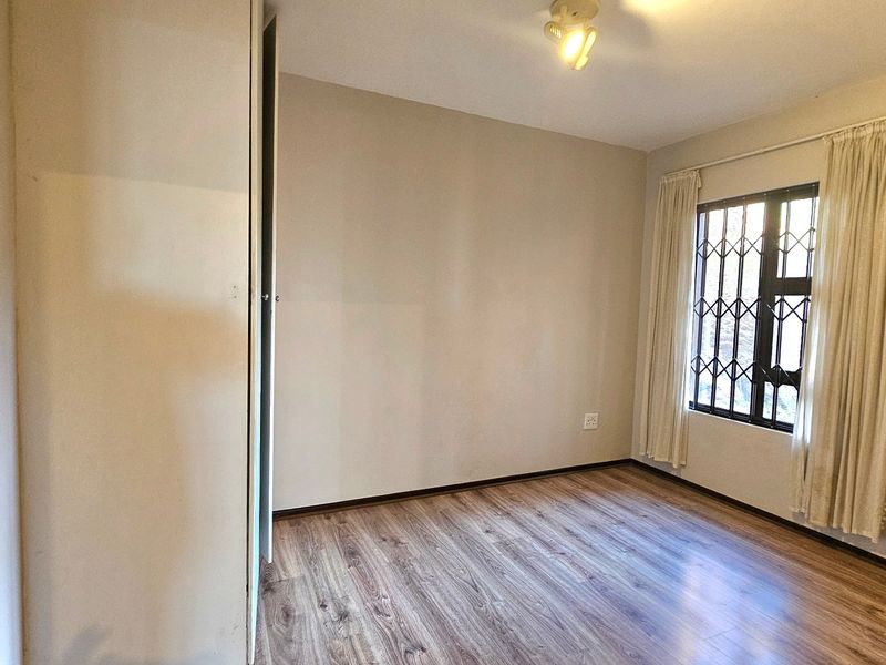 A bright, empty bedroom featuring modern laminate flooring, a built-in cupboard, and a window fitted with a security gate.