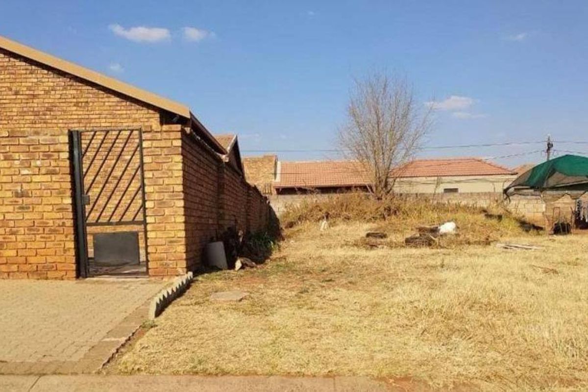 A view of the rear yard featuring a brick boundary wall, paved driveway area, and an unmaintained garden space.