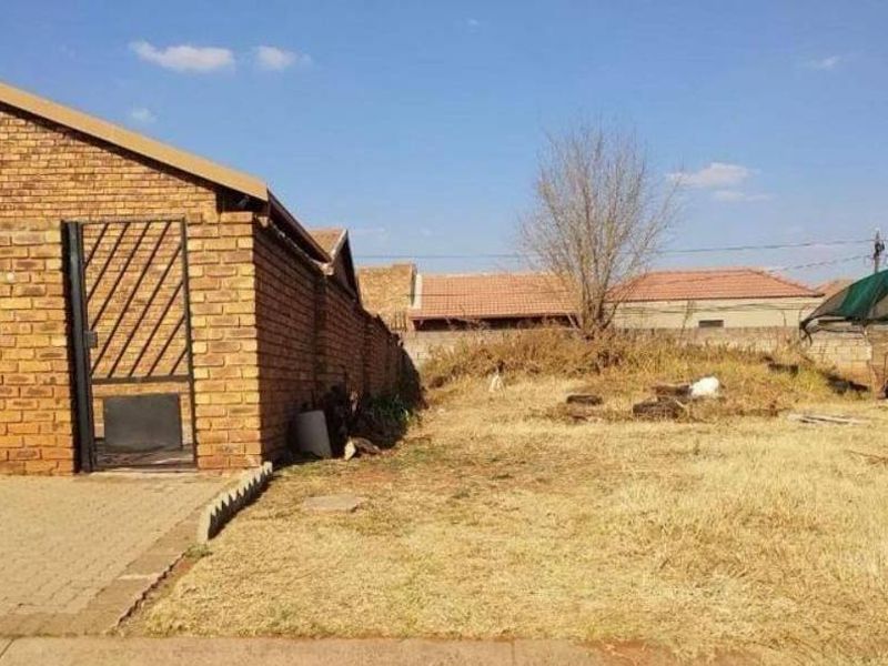 A view of the rear yard featuring a brick boundary wall, paved driveway area, and an unmaintained garden space.