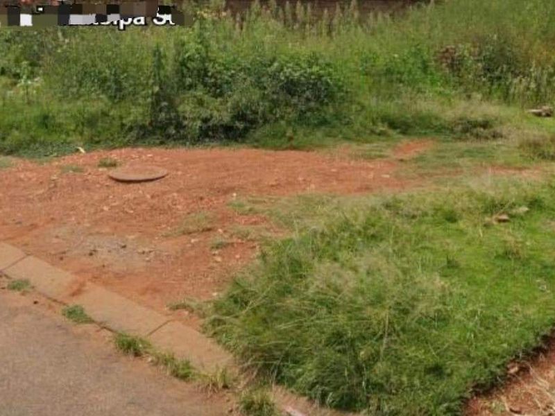 A view of a vacant residential plot showing natural ground, grass, and a manhole cover adjacent to the street kerbing.