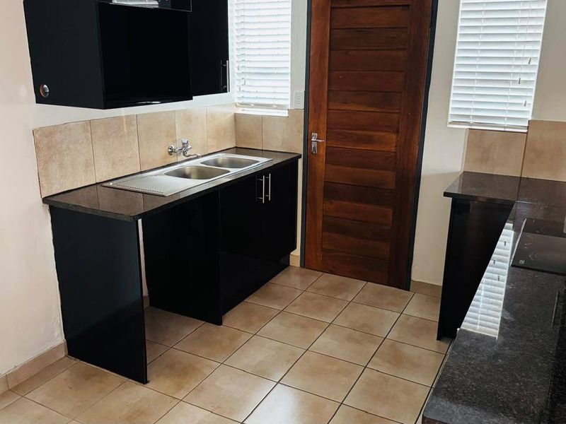 A functional kitchen featuring dark cabinetry, granite countertops, a double bowl sink, and neutral floor tiling with natural light from window blinds.