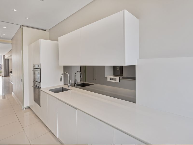 Modern minimalist kitchen featuring sleek white handleless cabinetry, integrated appliances, a mirrored splashback, and polished tiled flooring in an open-plan apartment layout.