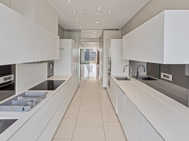 A modern, minimalist galley-style kitchen featuring sleek white cabinetry, quartz countertops, integrated appliances, and polished tile flooring in a bright, contemporary apartment.
