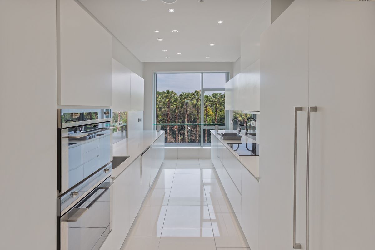 Modern galley-style kitchen featuring sleek white cabinetry, integrated appliances, stone countertops, and large windows with a scenic palm tree view.