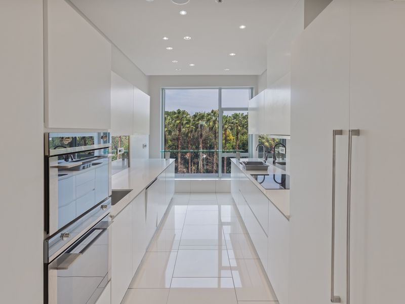 Modern galley-style kitchen featuring sleek white cabinetry, integrated appliances, stone countertops, and large windows with a scenic palm tree view.