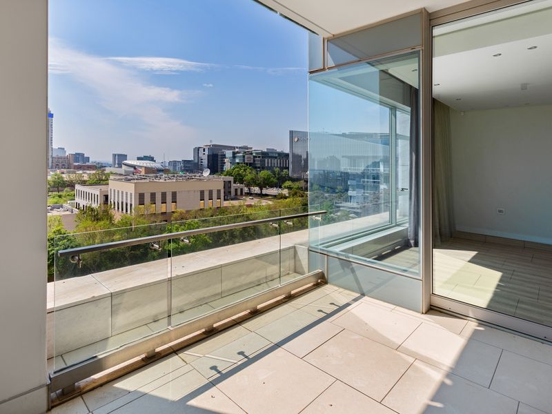 A modern, spacious balcony featuring a sleek glass balustrade, stone flooring, and expansive sliding glass doors offering panoramic city views.