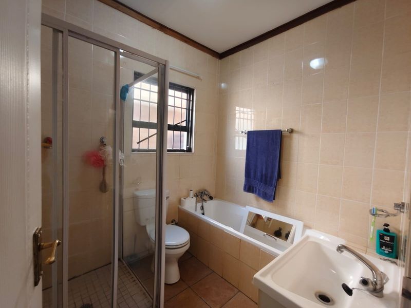 A functional bathroom featuring a shower enclosure, bathtub, toilet, and washbasin with neutral wall tiling and security bars on the window.