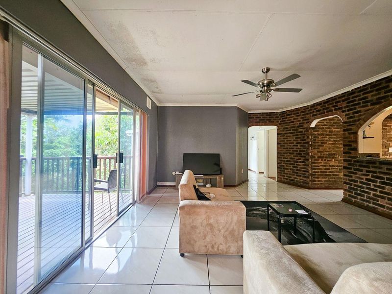 Spacious living room featuring tiled floors, an exposed brick feature wall, ceiling fan, and large sliding glass doors leading to an outdoor deck.