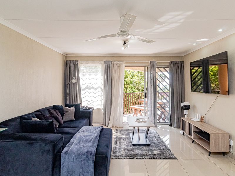 A bright and well-maintained living room featuring tiled flooring, a ceiling fan, and direct access to an outdoor balcony through sliding glass doors.