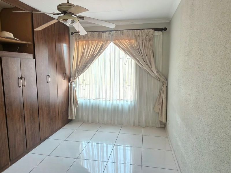 A clean, empty bedroom featuring built-in wooden cupboards, tiled flooring, and a ceiling fan, with natural light entering through a window with security bars.