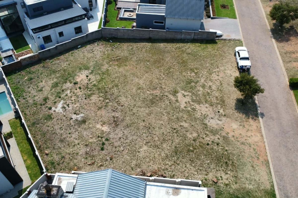 An aerial view of a vacant residential plot situated within a developed estate, featuring surrounding boundary walls and adjacent paved road access.