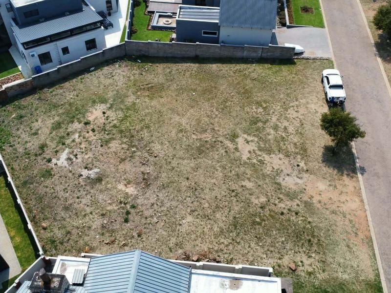 An aerial view of a vacant residential plot situated within a developed estate, featuring surrounding boundary walls and adjacent paved road access.