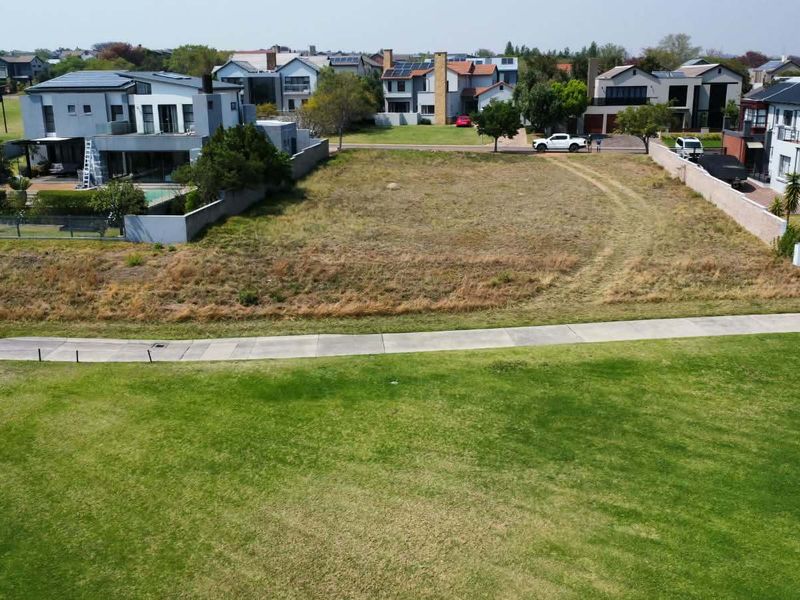 An aerial view of a vacant residential plot situated in a developed neighborhood, featuring a grass verge and paved walkway in the foreground.