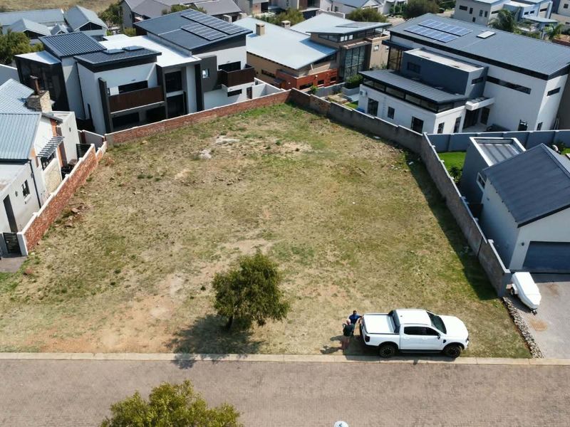 An aerial view of a vacant residential plot situated within a developed estate, featuring boundary walls and street access.