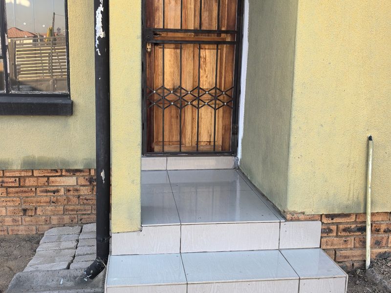Front entrance featuring a wooden door with a security gate, tiled steps, and brick facade detailing, showing signs of wear and unfinished exterior landscaping.