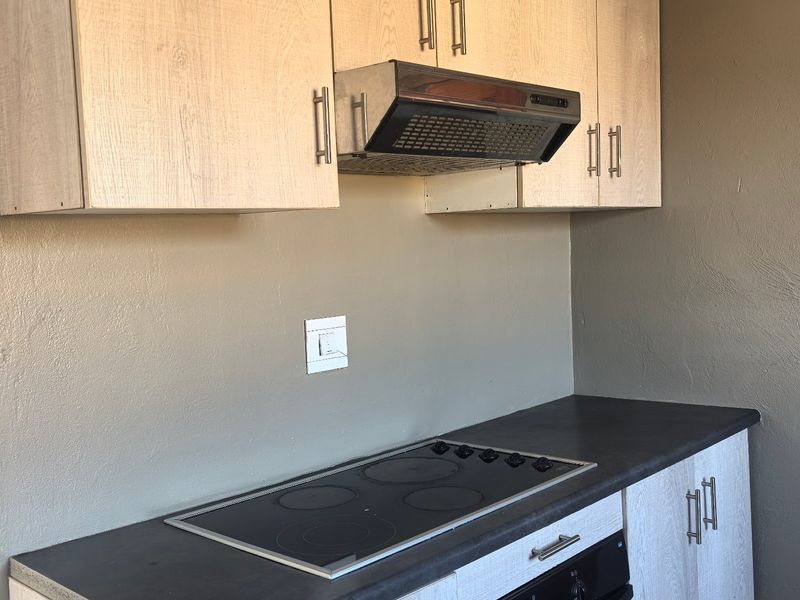 A compact kitchen area featuring light wood-grain cabinetry, dark laminate countertops, a built-in electric oven, and a hob with an overhead extractor fan.